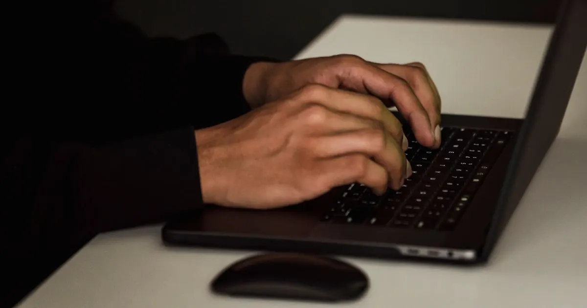 A hooded figure typing on a laptop in the dark